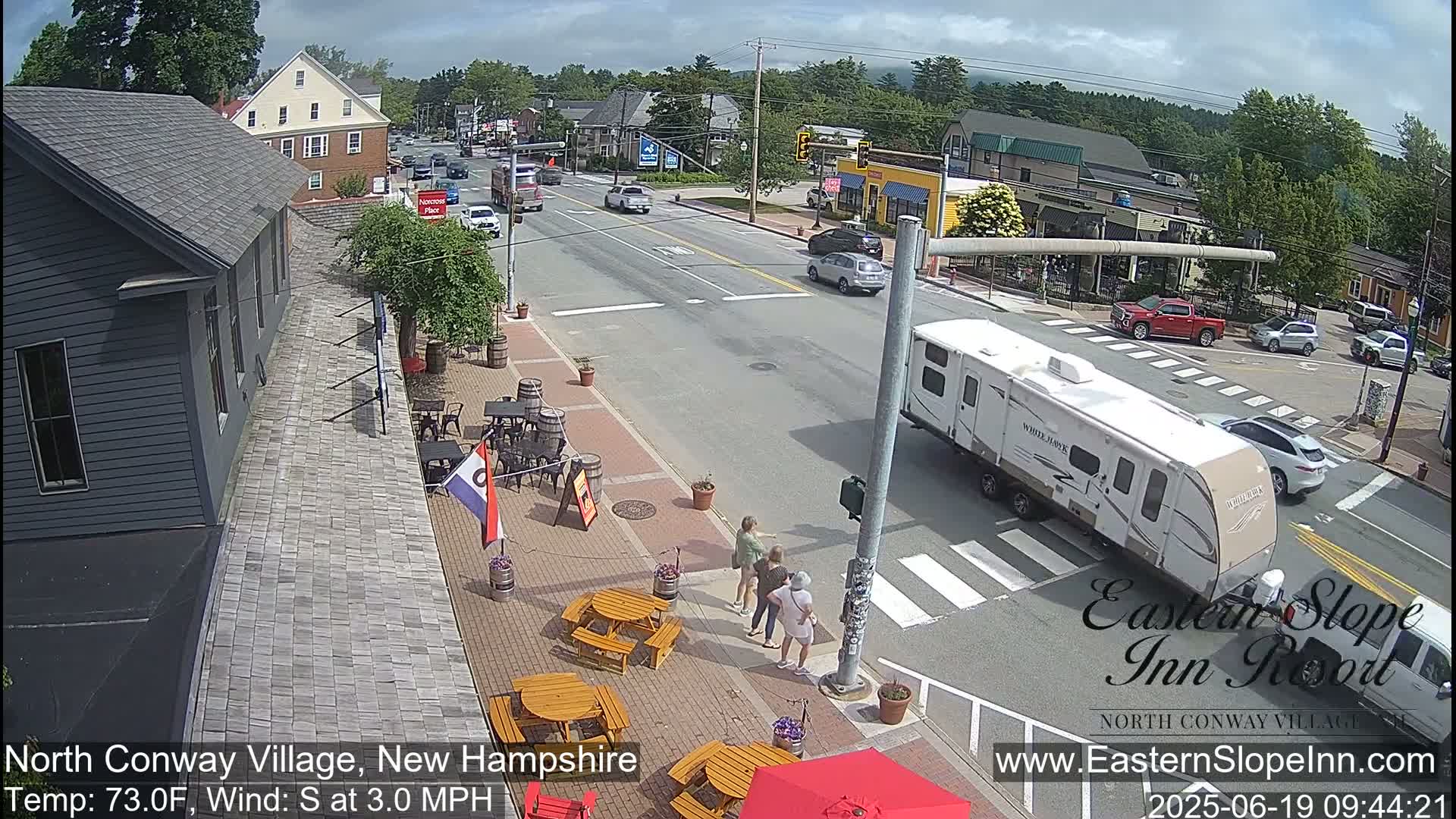 A partly sunny day shows a street scene with cars and a camper, and people standing near outdoor tables at a restaurant.