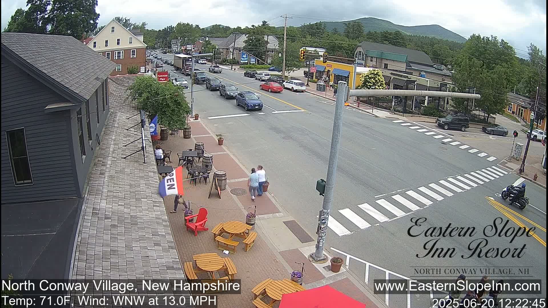 A street scene in a village shows cars driving on a road that intersects with a crosswalk, pedestrians walking on the sidewalk, and outdoor seating in front of a building under partly cloudy skies.