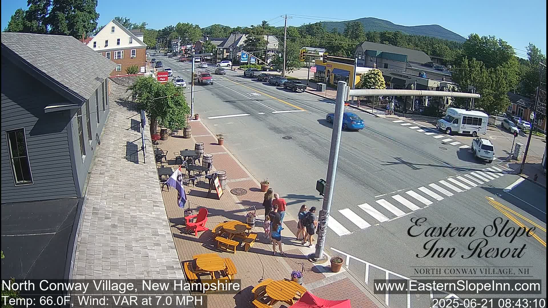 A sunny street scene shows a village intersection with pedestrians near outdoor seating at a restaurant, cars on the road, and a partly cloudy sky with mountains in the distance.