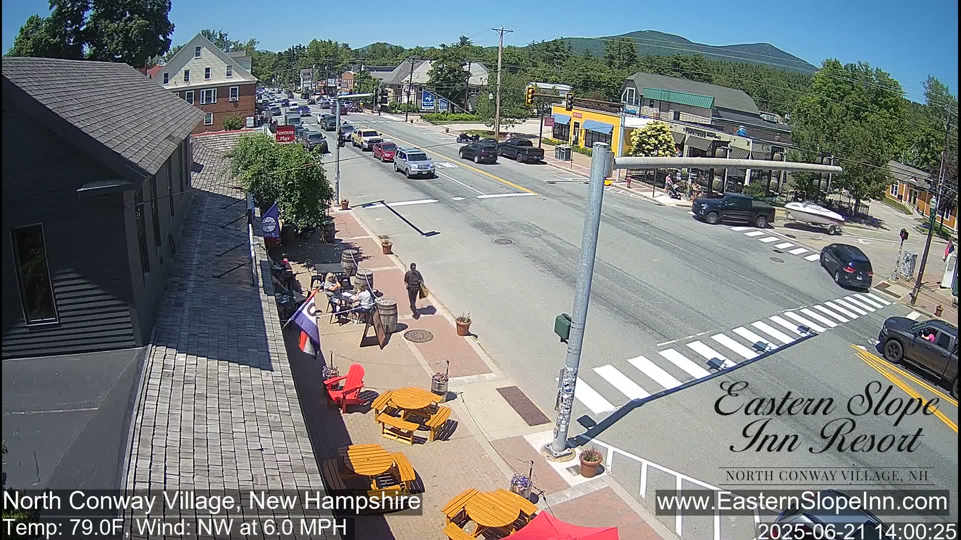 A sunny street scene shows several cars driving on a road that intersects with another road, with outdoor seating and pedestrians near buildings.