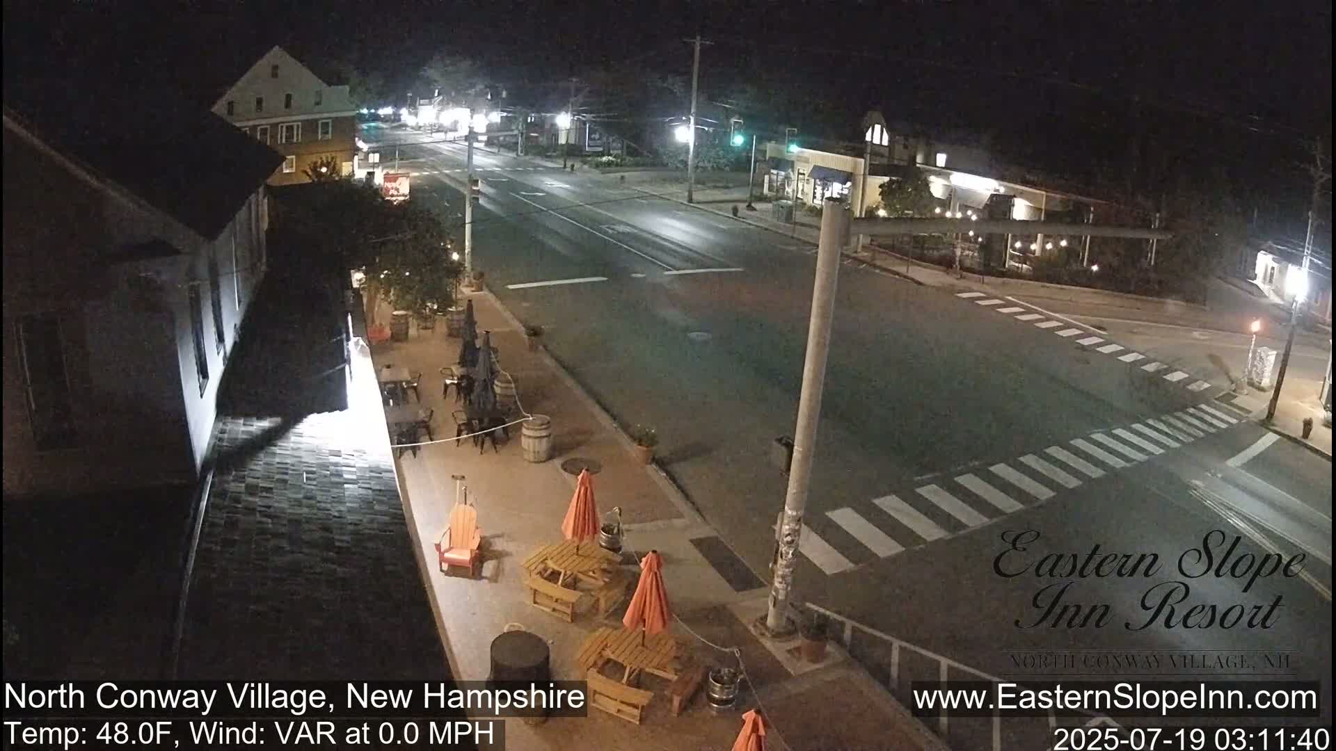 A nighttime view of a mostly empty street intersection in a town, with outdoor seating areas at a nearby building under mostly clear skies.
