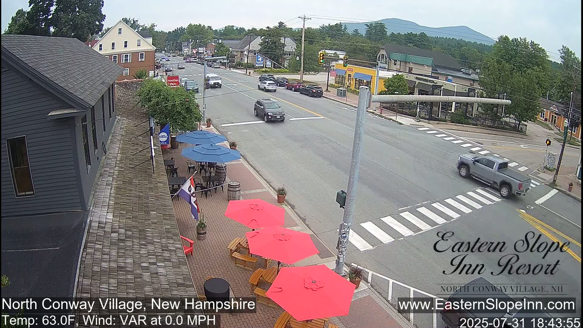 A street scene on a sunny day shows a road intersection with several cars, a sidewalk with red and blue patio umbrellas and outdoor seating, and buildings in the background.