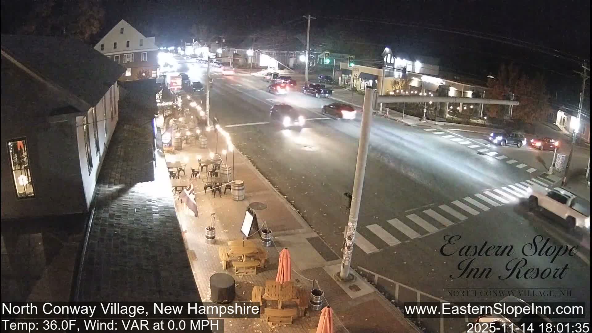 A clear night in a small village reveals a well-lit main street with several cars driving, their headlights glowing, bordered by an outdoor dining area featuring tables, chairs, and decorative barrels beside a multi-story building.