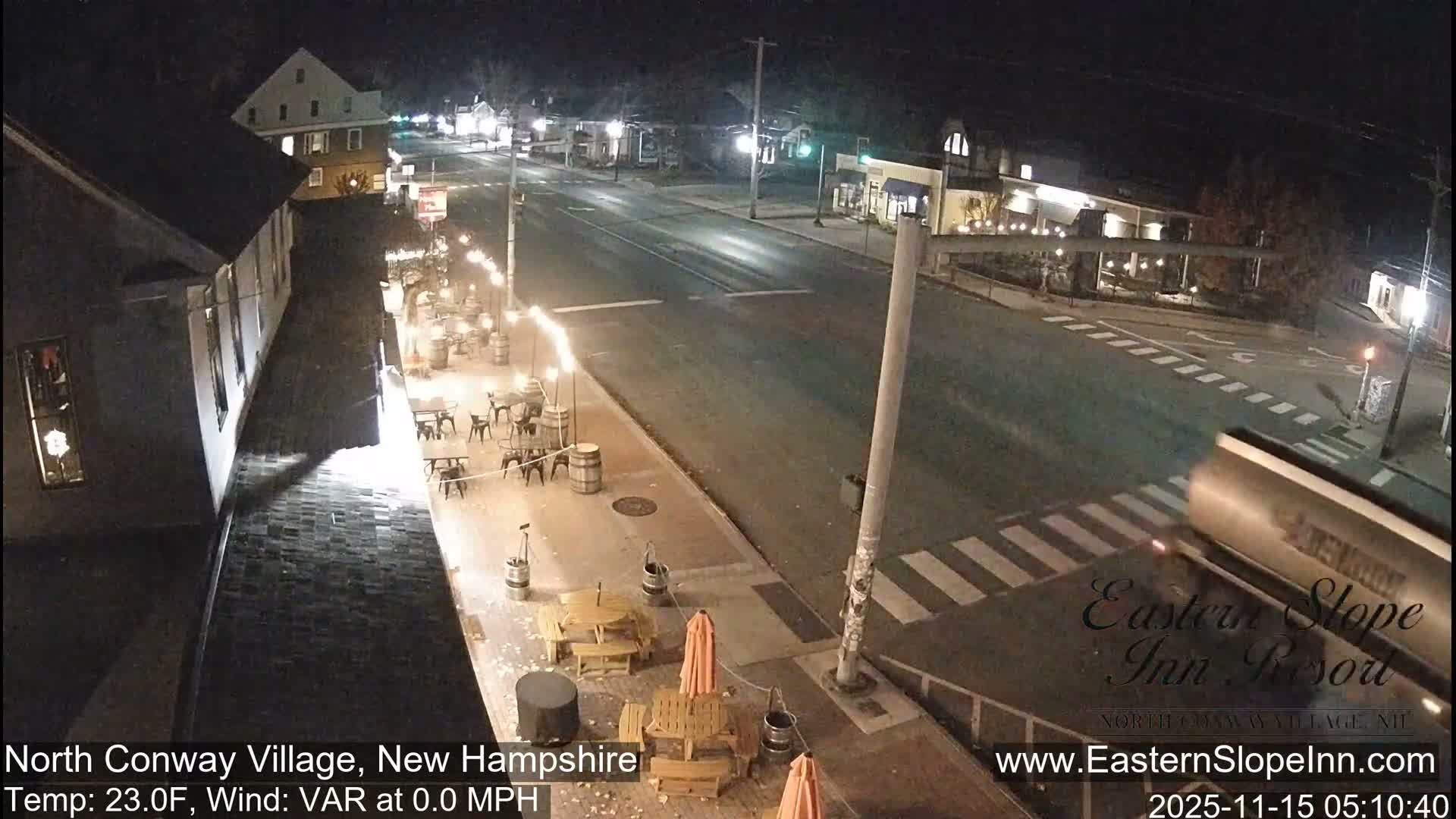 At night, a quiet village street is illuminated by building windows and streetlights, revealing outdoor dining areas with string lights and tables, with a blurry vehicle passing a crosswalk under clear, cold, and calm conditions.
