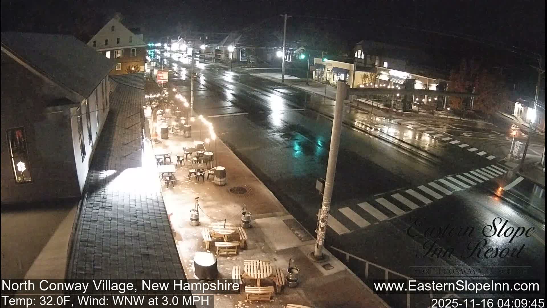 A nighttime view reveals a wet village street lined with illuminated buildings and outdoor patio seating, with reflective asphalt suggesting recent rain or damp conditions.