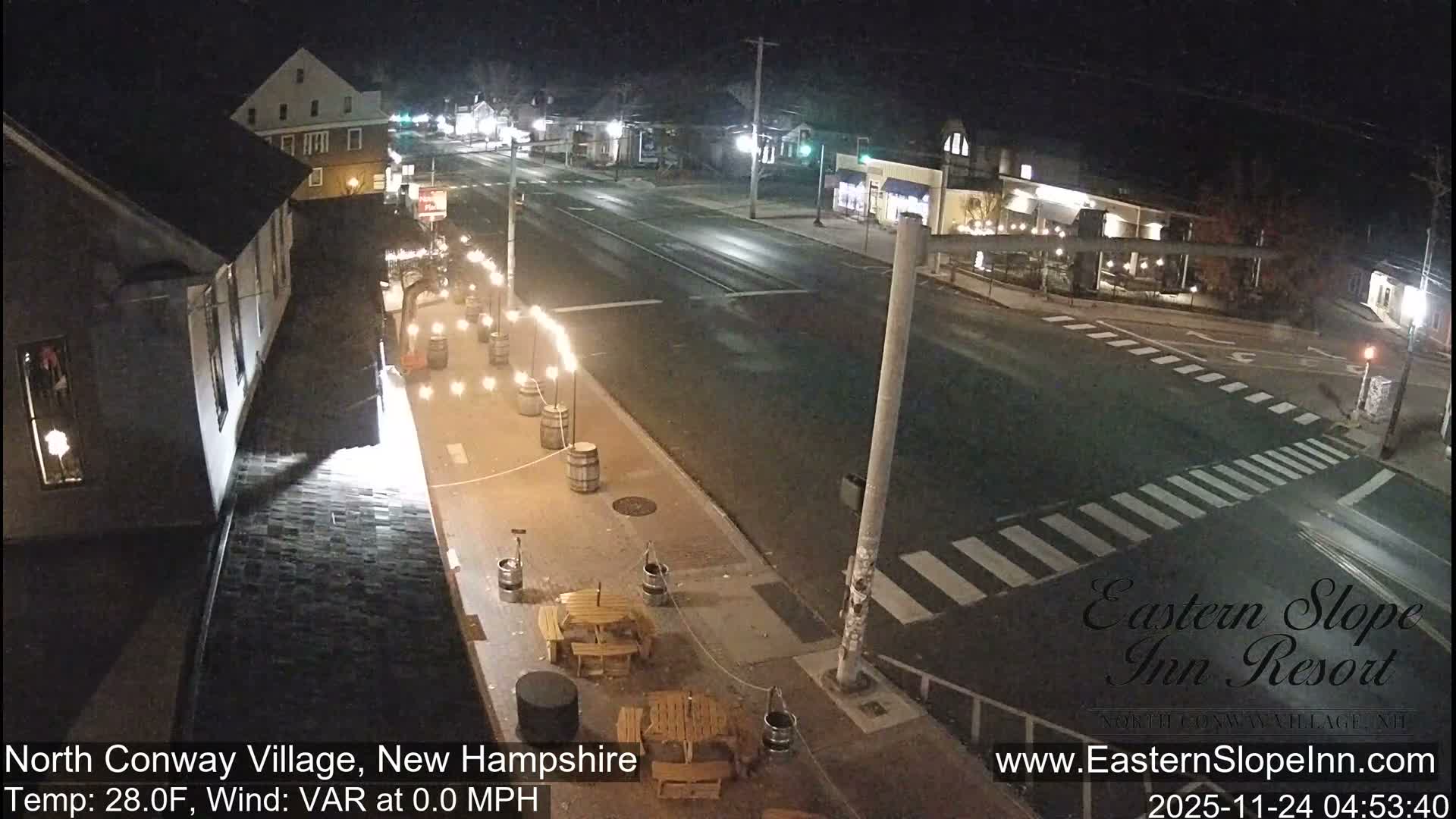 A dark nighttime view shows a quiet street lined with illuminated buildings and streetlights, with string lights and picnic tables on the left sidewalk, and the road surface appearing wet.