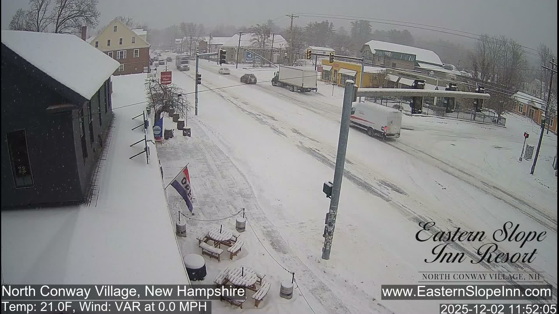 A snow-covered village street features buildings with snowy roofs, bare trees, and several vehicles navigating a slushy road, all under an overcast sky with light snow actively falling.