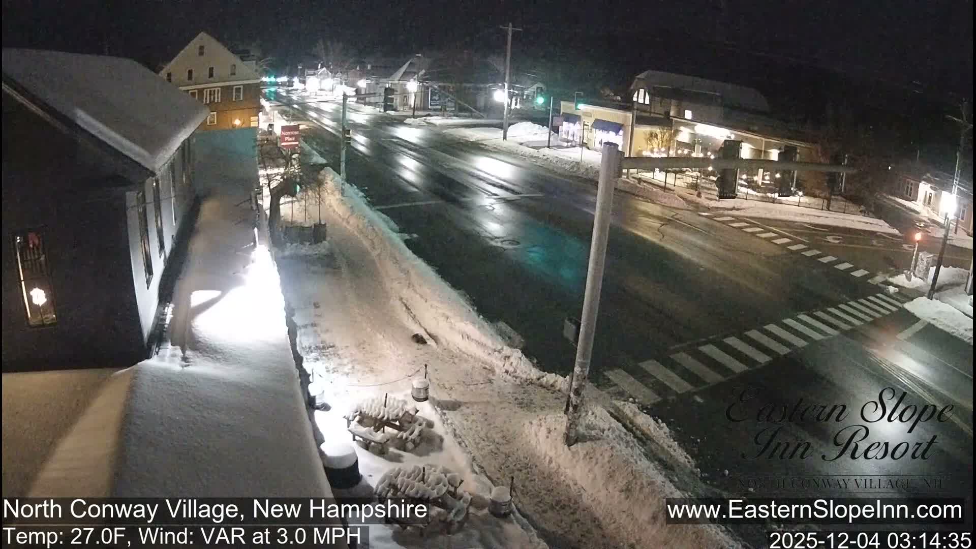 A quiet, snow-covered village street is illuminated at night under cold, wintery conditions, showcasing a reflective main road, clear crosswalks, and buildings with snow-dusted roofs and sidewalks.