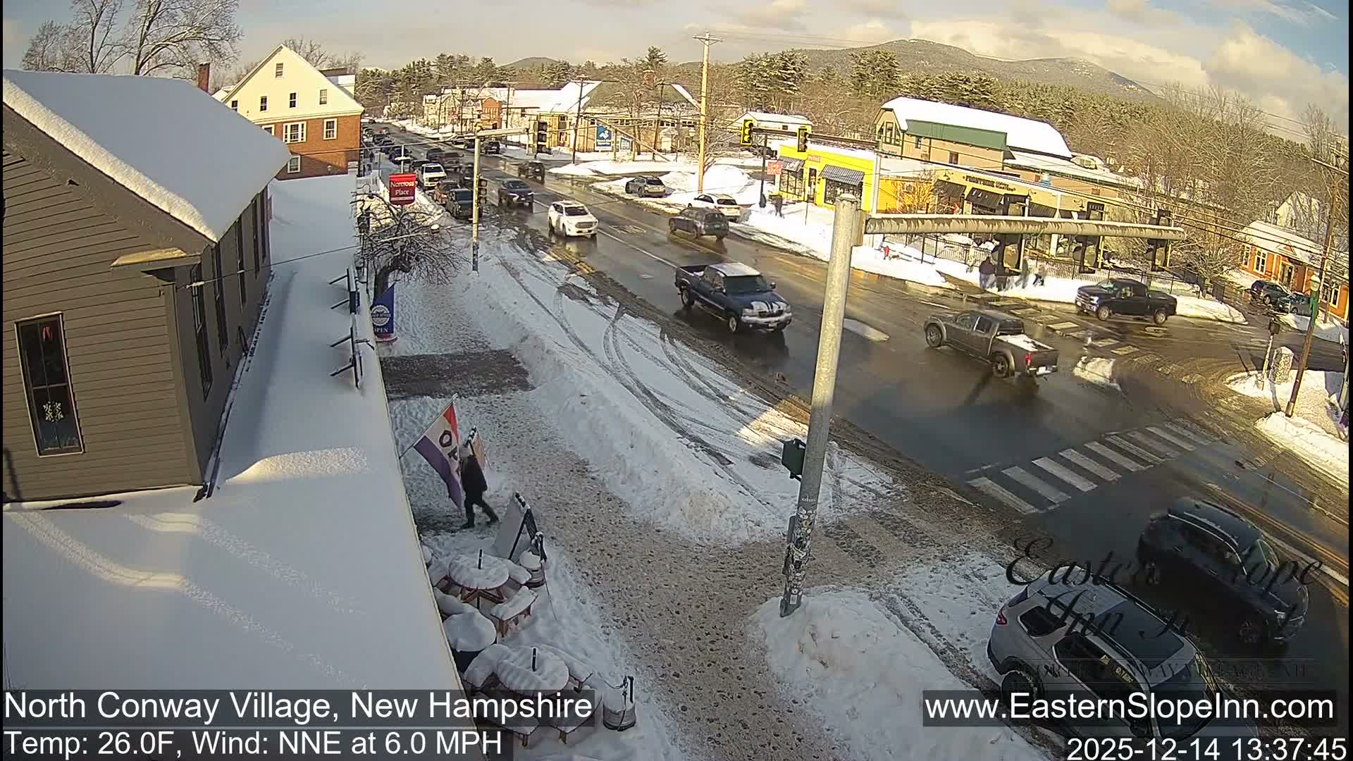 A quiet, snow-covered village street is illuminated at night under cold, wintery conditions, showcasing a reflective main road, clear crosswalks, and buildings with snow-dusted roofs and sidewalks.