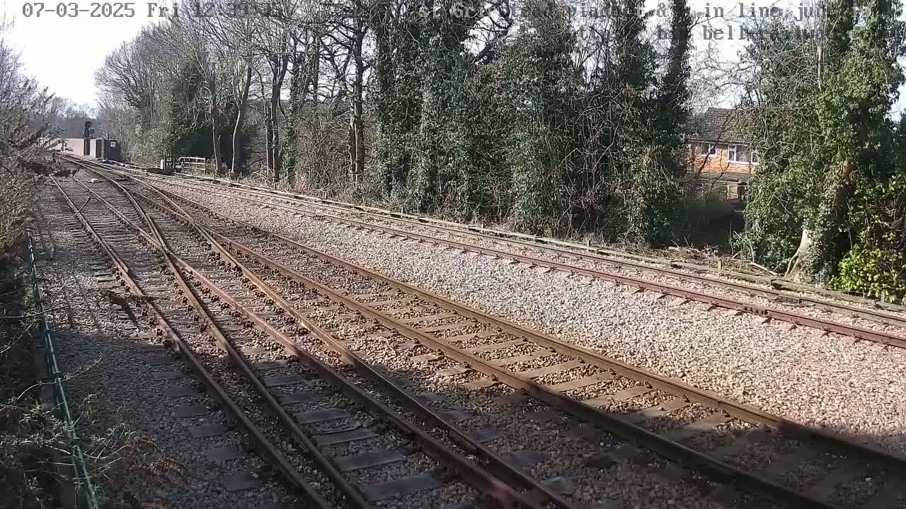 A sunny day view of a railway track splitting into multiple directions, bordered by trees and shrubs, with a building visible in the distance.