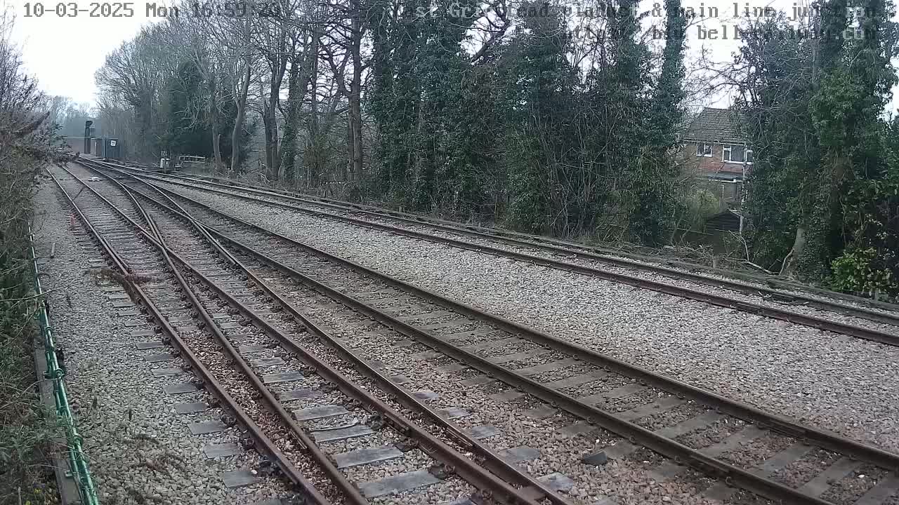 Multiple train tracks diverge amidst a landscape of trees and a residential building under an overcast sky.