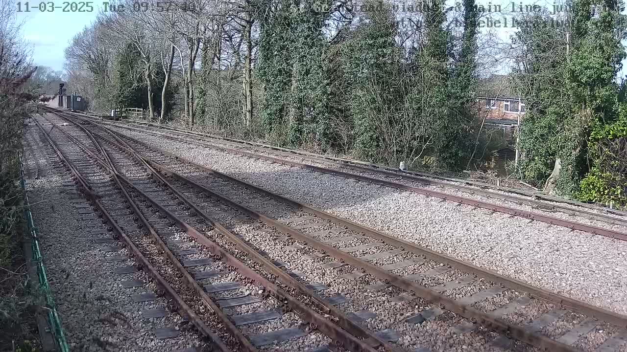 A railway track with a point splits into two directions under a sunny sky, passing through a wooded area with a house visible in the background.