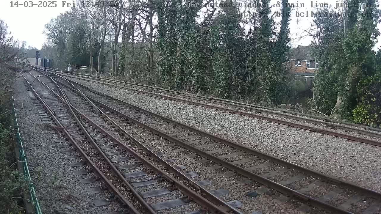 A view of railway tracks diverging on a cloudy day, with trees and a building visible in the background.