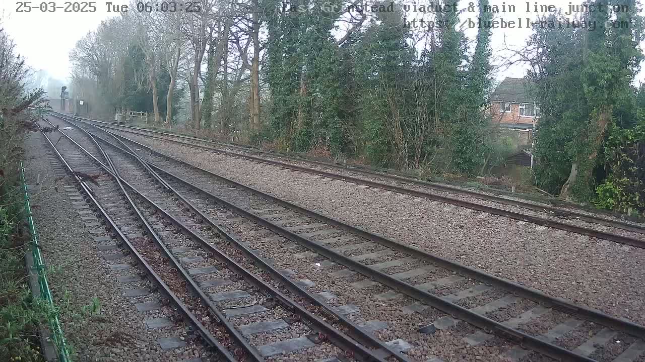 A view of a railway track junction on an overcast day, bordered by trees and a house visible in the distance.