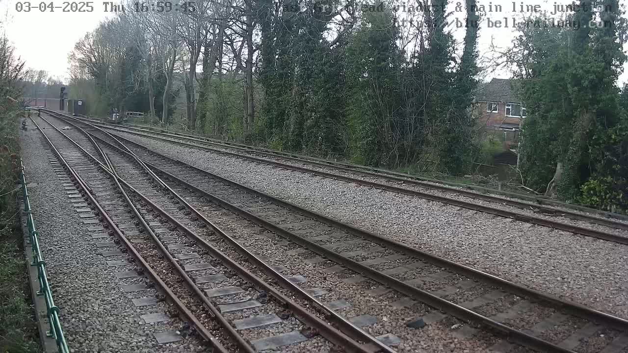 Converging train tracks on a gravel bed, bordered by trees and a house in the distance, under an overcast sky.