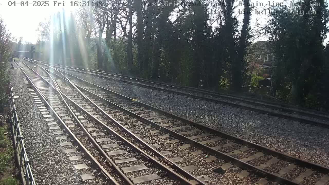 A sunny day shows a view of railway tracks branching off to the left, bordered by trees and a stone wall.