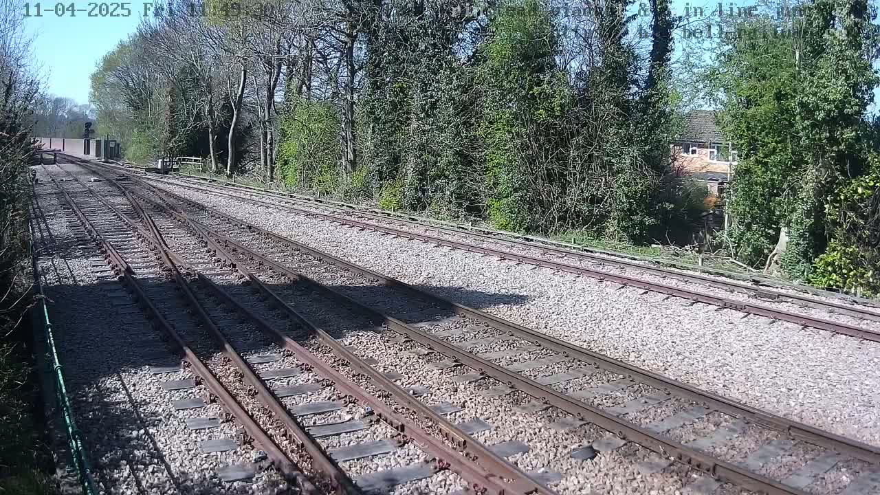Multiple train tracks extend into the distance on a sunny day, bordered by lush green vegetation and a glimpse of a building in the background.