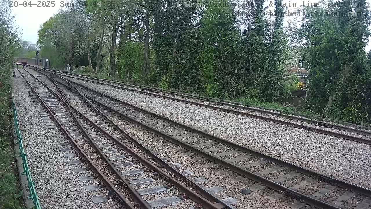 Several train tracks diverge through a gravel path bordered by lush green trees and shrubbery on a partly cloudy day.