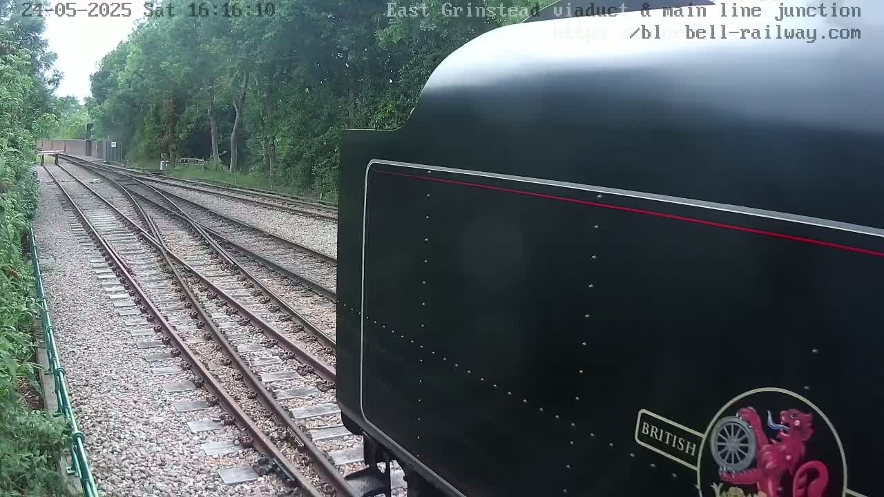 A dark-colored train car is seen from the side, approaching a railway junction with multiple tracks under an overcast sky.