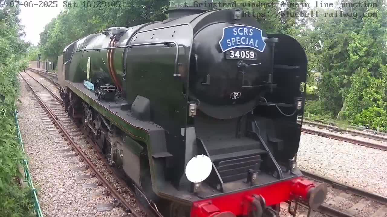 A dark-green steam locomotive travels along a railway track, surrounded by lush green trees under partly cloudy skies.