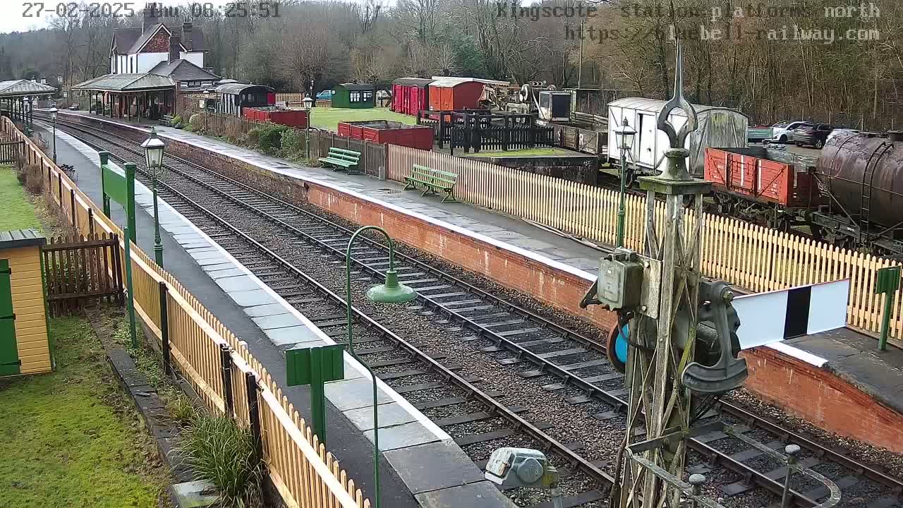 A train station platform with multiple railway tracks, several railway cars, and a station building is visible, under an overcast sky.