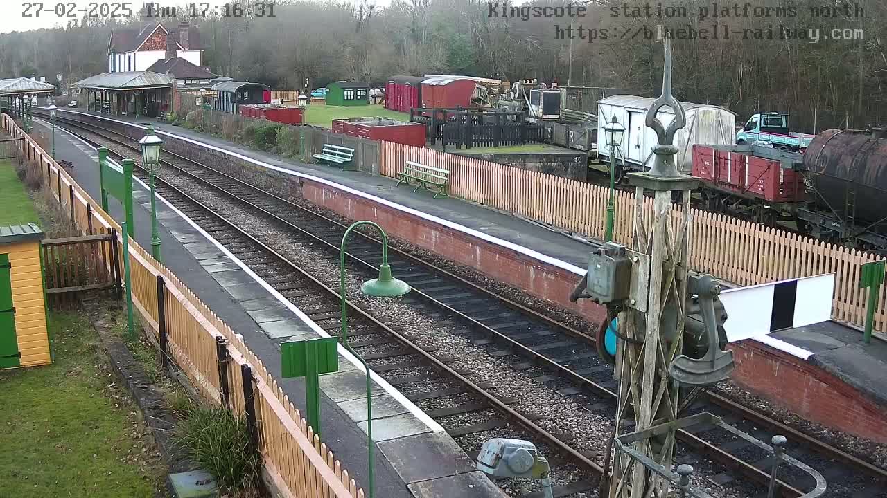 A railway station platform with two sets of tracks, surrounded by wooden fences and various railway cars, on an overcast day.