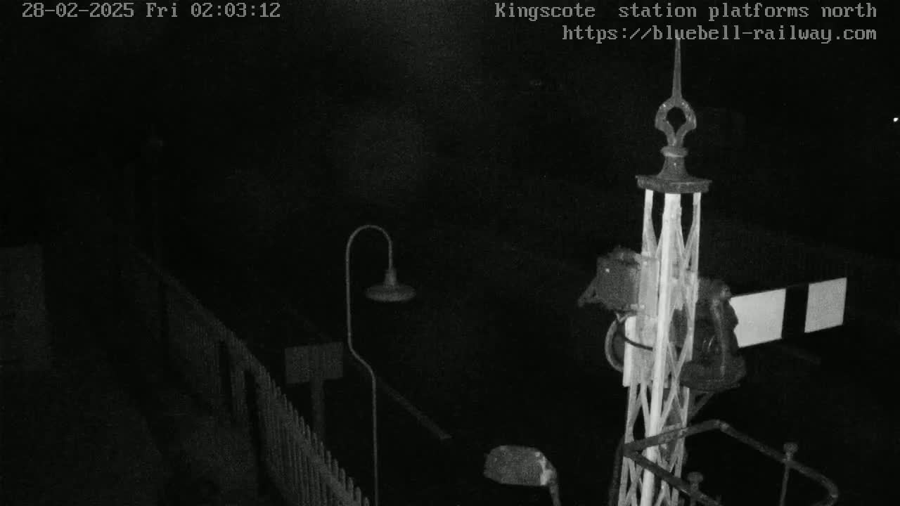 A nighttime, low-light view of a railway signal and platform area with a fence and lamppost.
