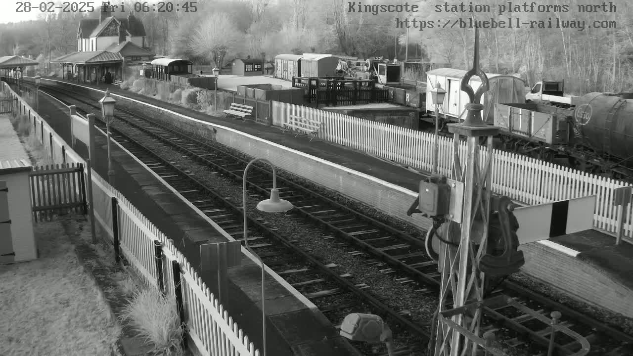 A black and white, overhead view of a train station platform with railway tracks, train cars, and buildings on a cold, possibly snowy or icy, day.