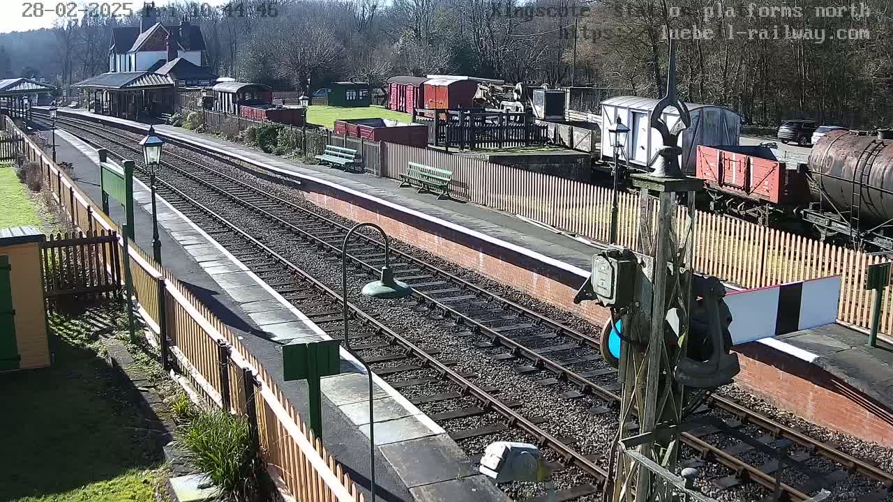 A sunny day at a train station with multiple railway tracks, various train cars, and a station building.