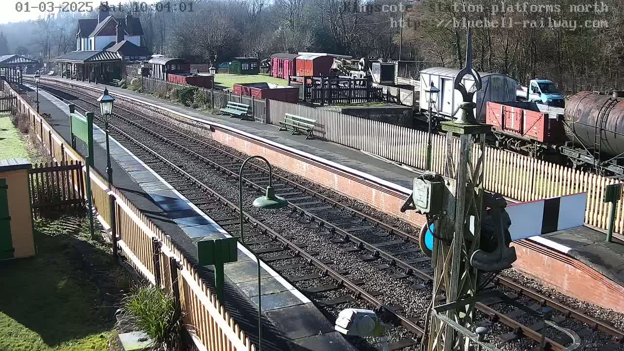 A sunny day at a train station with multiple train cars and a station building visible, alongside train tracks extending into the distance.