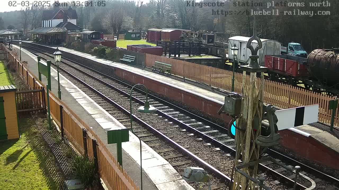 A sunny day at a railway station with multiple train cars and a platform, surrounded by wooden fences and greenery.