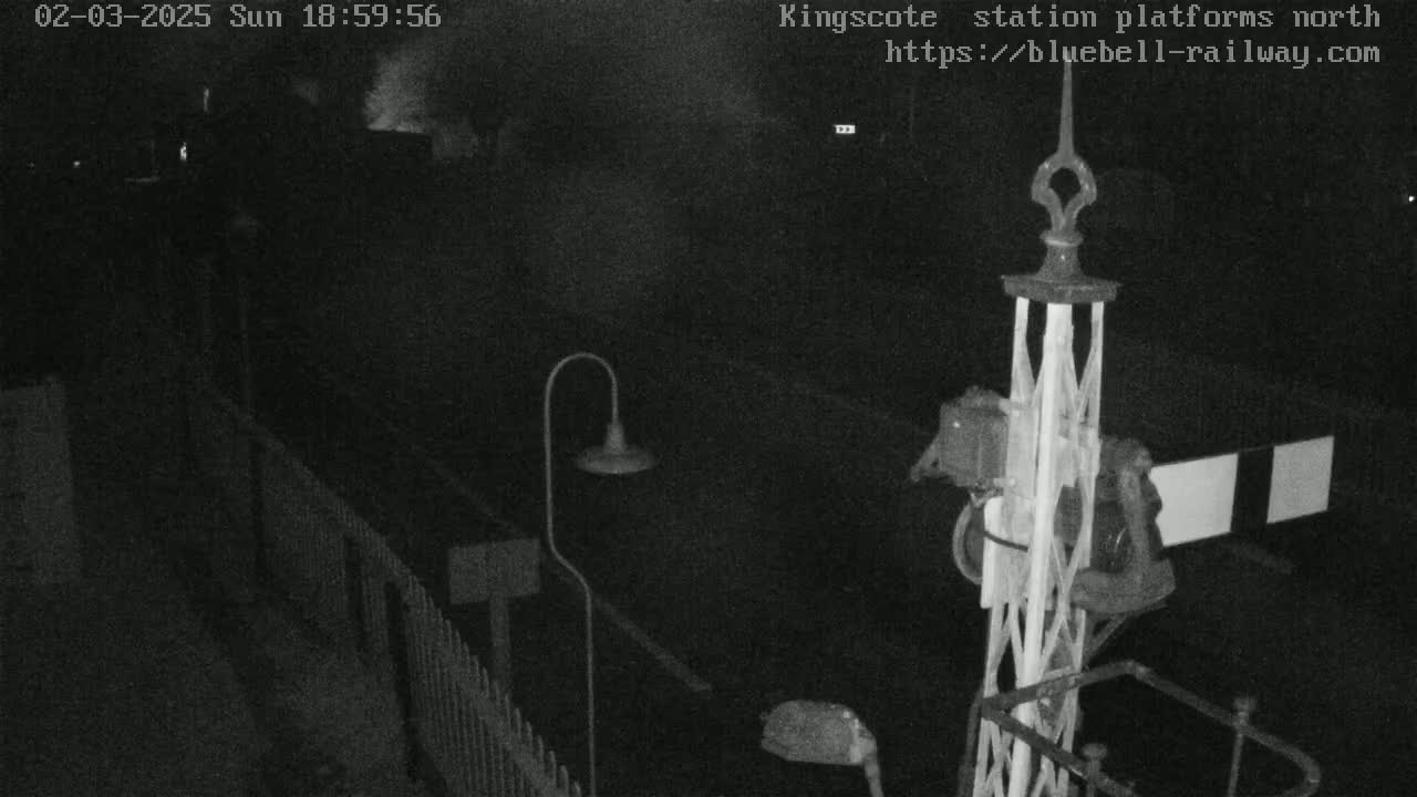A nighttime, low-light view of a train station platform, showing a metal signal post and a portion of a fence.