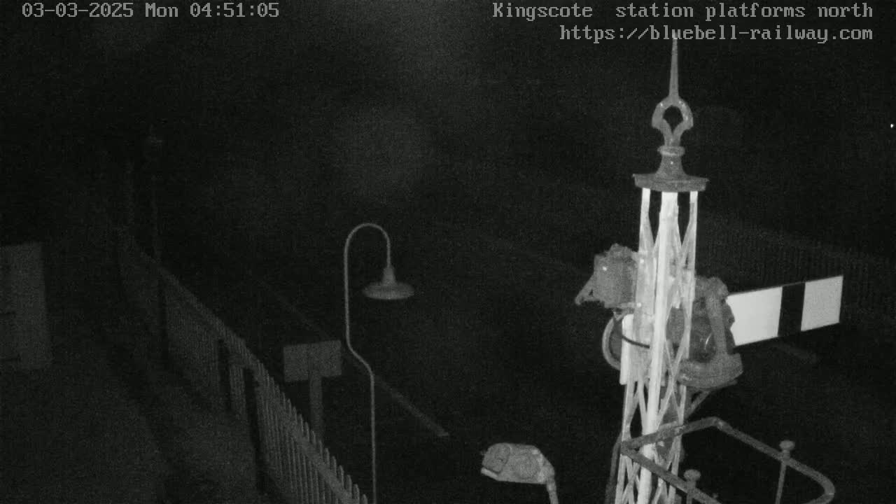 A nighttime, low-light view of a railway signal and platform area, featuring a tall metal signal post and a fence.