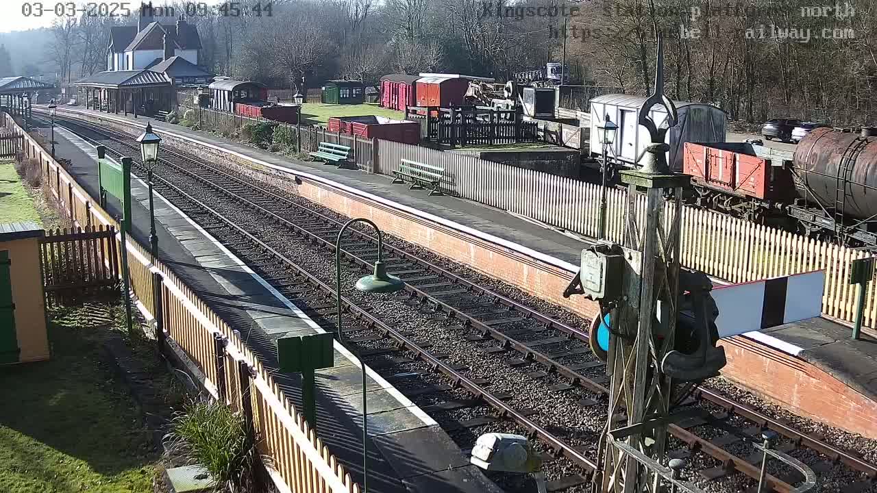 A sunny day at a train station shows multiple railway tracks, a platform with benches, and various railway cars and buildings.