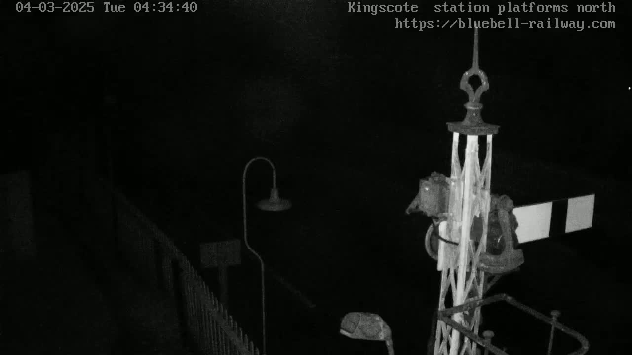 A nighttime, low-light image shows a metal railway signal and lamppost, partially obscured by darkness, near a fence.