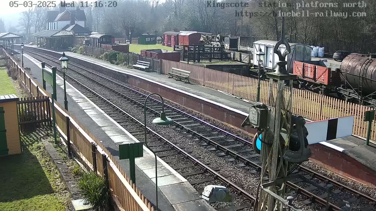 A sunny day at a train station with multiple railway tracks, several train cars, and a platform with wooden fences.