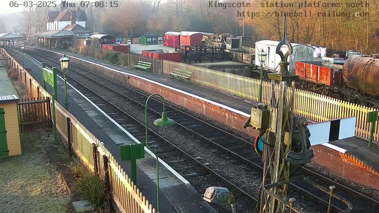 A sunny view of a train station platform with railway tracks, passenger shelters, and several freight cars visible.