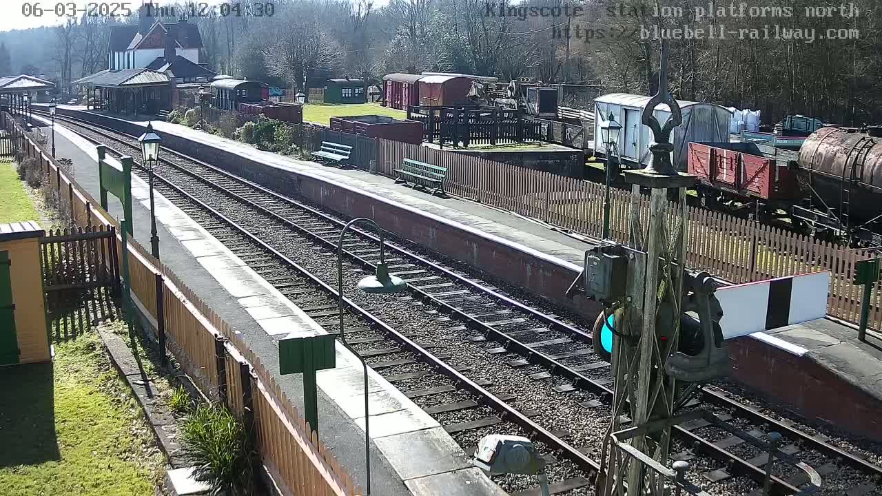 A sunny day at a train station shows a railway platform with tracks, railcars, and a station building.