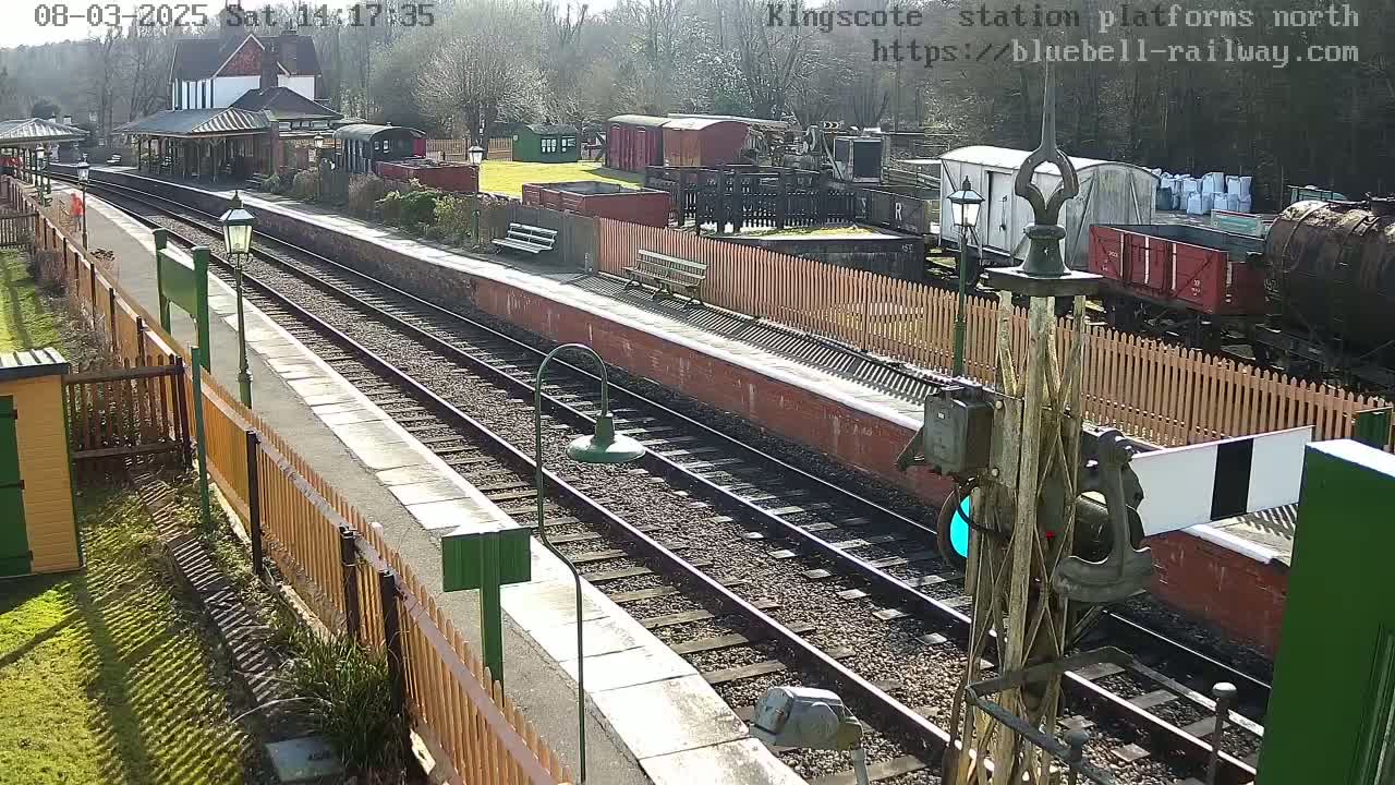 A sunny day at a train station with two sets of tracks, a platform, and several rail cars and buildings visible.