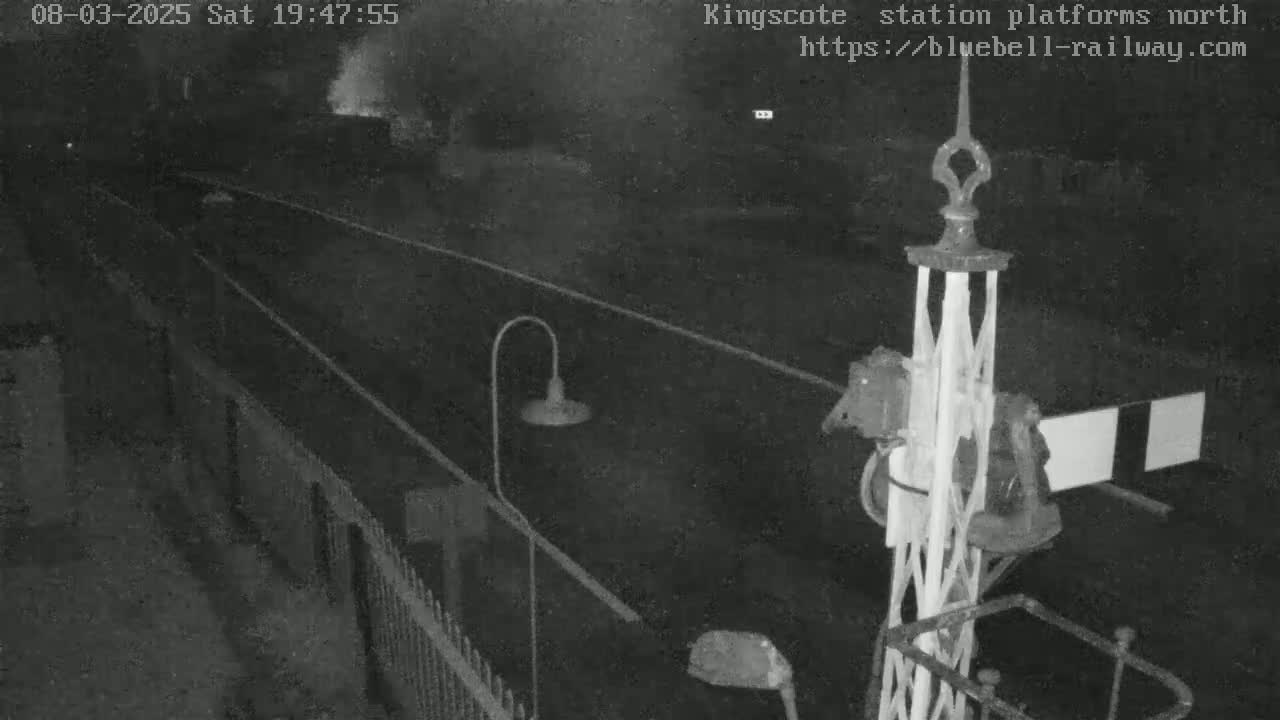 A nighttime view of a railway platform with a signal and a fence, under dark conditions.