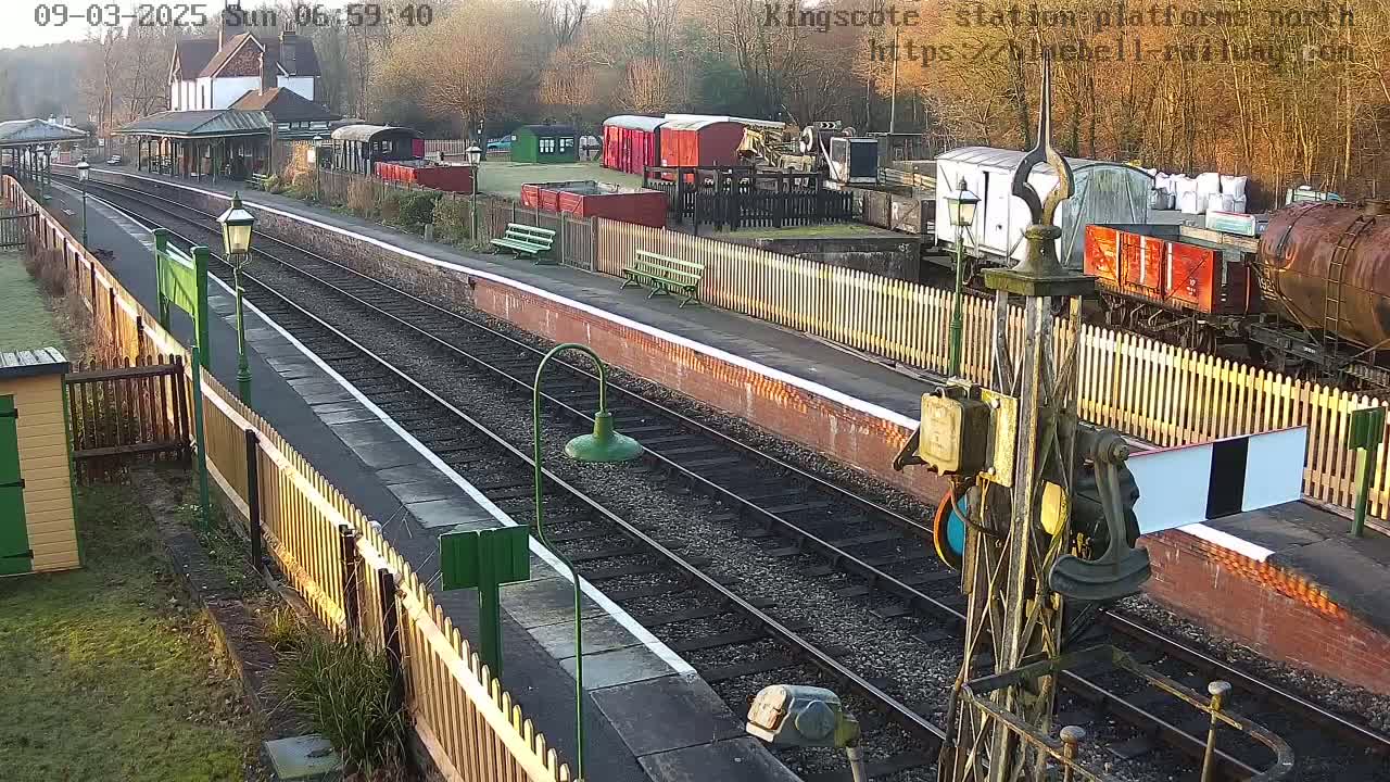 A sunny view of a train station platform with multiple train cars and railway tracks, surrounded by wooden fences and buildings.