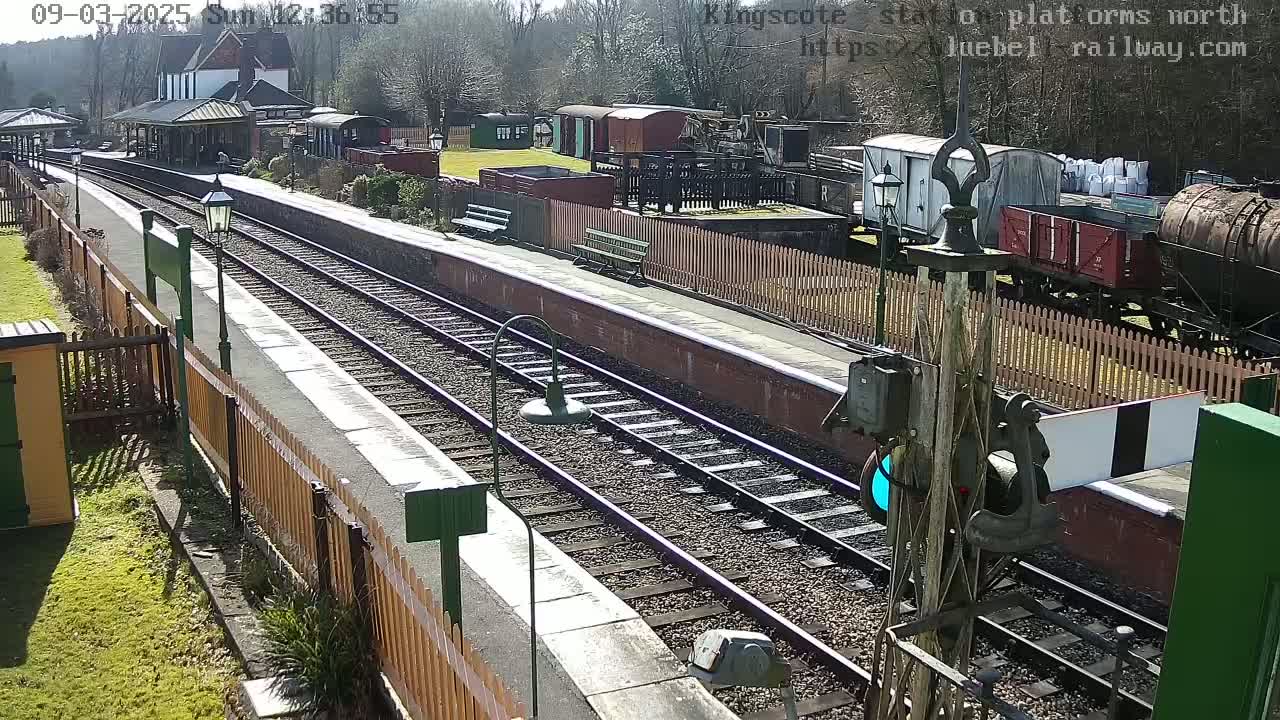 A sunny day at a train station with railway tracks extending into the distance, featuring various train cars and a station building.