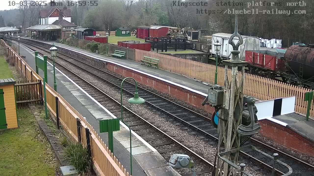 An overcast day at a train station with multiple train cars and a station building visible, and two sets of train tracks extending into the distance.