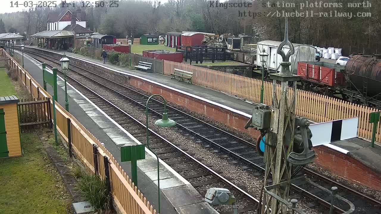 A view of a train station platform with multiple train tracks, railway cars, and a person walking in the distance, on an overcast day.