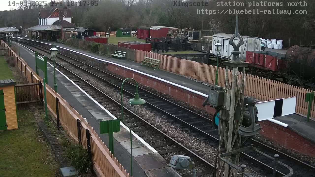A railway station platform with two parallel tracks, several railway cars, and a station building in the background, on an overcast day.