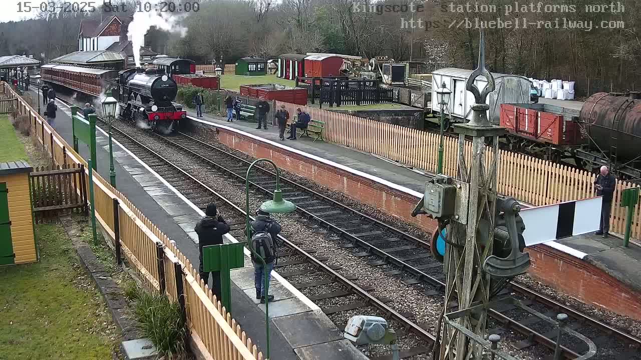 A steam train pulls into a railway station platform on an overcast day, with several people standing and watching nearby.