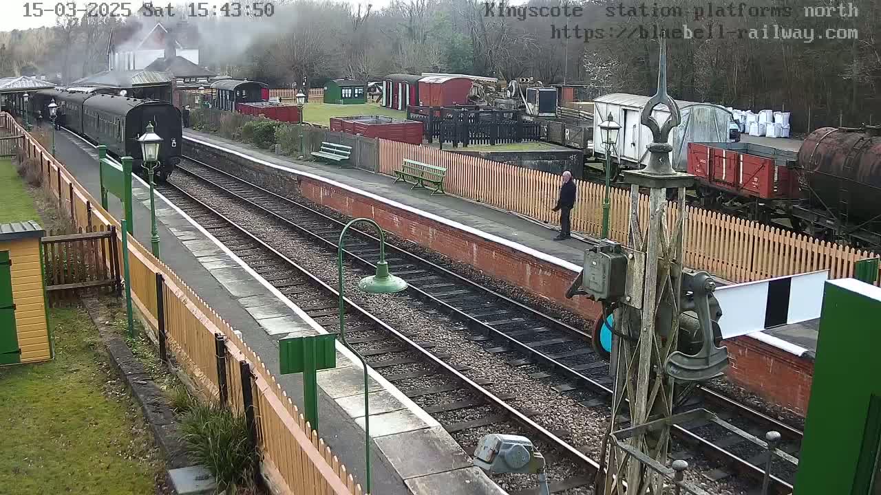 A steam train is arriving at a railway station platform on a cloudy day, with various railway cars and buildings visible in the background.