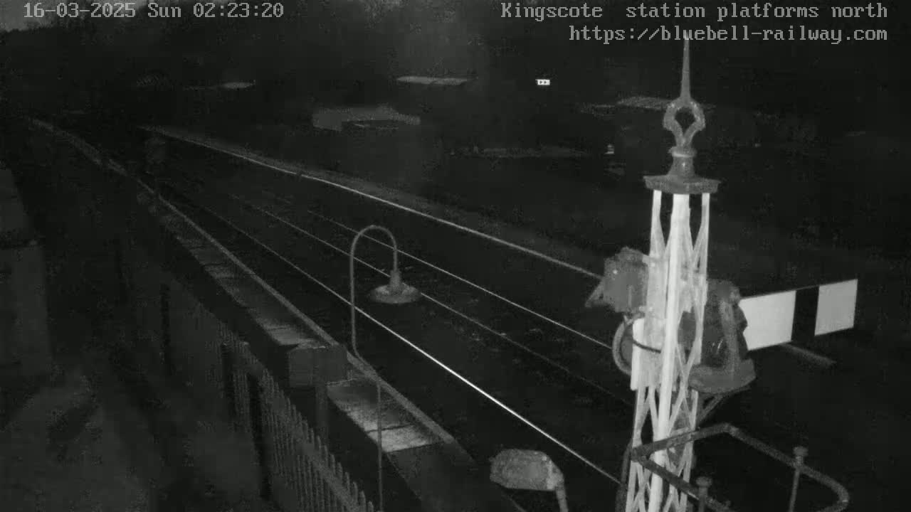 A nighttime, low-light view of empty train tracks at a station platform, with a metal signal post visible in the foreground.