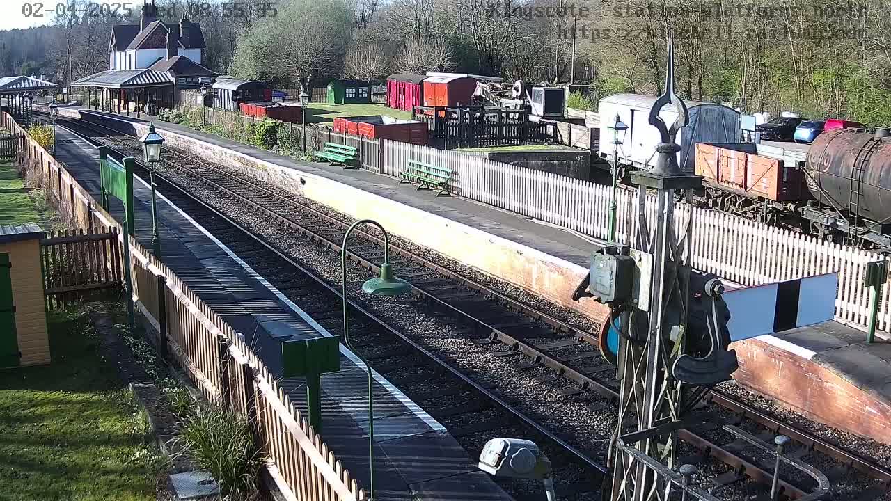 A sunny day at a train station with railway tracks, a platform, several freight cars, and a building in the background.