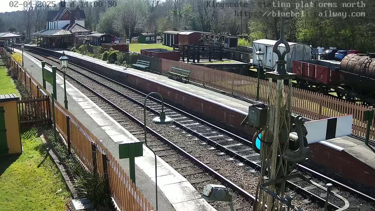 A sunny day at a railway station with two parallel train tracks, various railway cars, and a station building in the background.